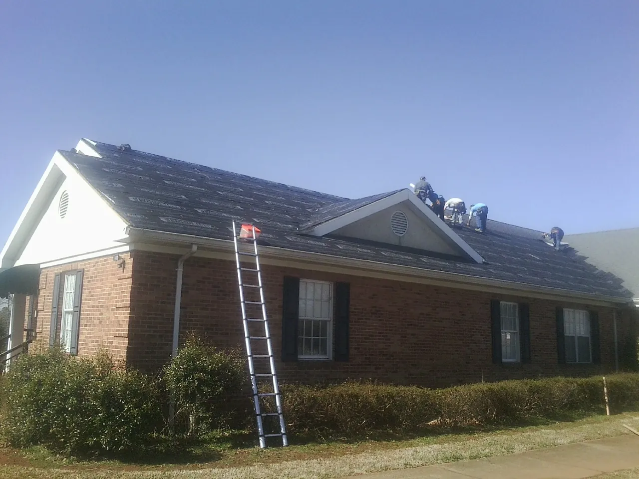 Crew tearing shingles off a brick ranch roof with a ladder set — Metal Roof Repair work by our crew in Langlois