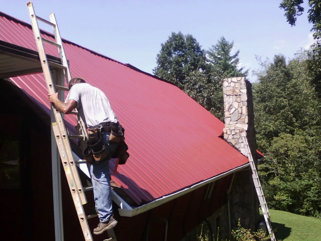 Roofer on a ladder beside a red metal roof and stone chimney — Soffit & Fascia Repair by Roofing Brothers in Langlois